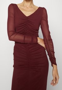 Burgundské šaty s dlouhými průhlednými rukávy; mají řasení po celé délce těla a sukně, výstřih do V, přiléhavý silueta. Hladká textura látky.