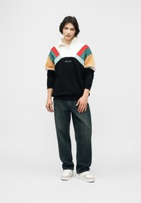 Νεαρό άτομο στέκεται με casual ντύσιμο, φορώντας πολύχρωμο φούτερ με φερμουάρ στη μέση, χαλαρά σκούρα τζιν και λευκά αθλητικά παπούτσια, σε απλό φόντο.