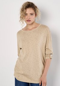 Beige långärmad skjorta med lös passform, mjuk textur, rullade ärmslut som är säkrade med knappar och rund halsringning. Subtilt mönster genom hela skjortan.
