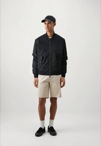 Dark navy bomber jacket with zip closure, side pockets, and ribbed cuffs, paired with beige shorts and black slip-on shoes. Light socks worn.