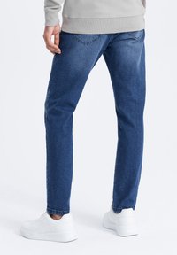 Siniset farkut suoran lahkeensuunnittelun kanssa, vähäisellä kulumisella, takataskuilla ja valkoisten tennareiden kanssa. Rakenteellinen kangas ja rento istuvuus.