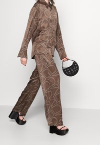 Henkilö kävelee päällään ruskea kuvioitu silkkipaita ja housut, pitäen kädessään pientä mustaa paljettilaukku, ja hänellä on mustat korokepohjaiset sandaalit.