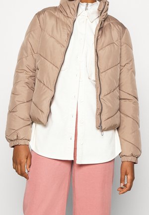 Person wearing a beige quilted zip-up jacket over a white buttoned shirt and pink pants, standing against a plain background.