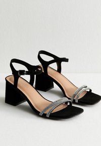 Sandali neri in suede con tacco, con punta aperta, caratterizzati da due cinturini decorati con cristalli e un tacco a blocco. Inclusa una cinghia regolabile alla caviglia.