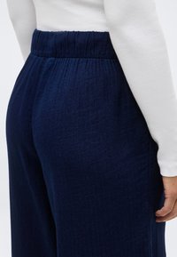 Nauwsluitende broek met elastische tailleband in marineblauw, gedragen met een wit, lang mouwig shirt, met zijkant- en achterkantdetails zichtbaar op een staand persoon.