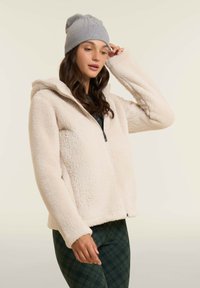 Giacca a felpa con cappuccio in morbido crema con chiusura a zip, caratterizzata da una vestibilità comoda e una superficie texturizzata; abbinata a un berretto in maglia grigia.