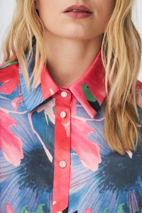 Kukkakuosiinen pusero, jossa eloisat pinkki, sininen ja vihreä värit. Punainen kaulus ja koristellut napit kaula-aukossa.