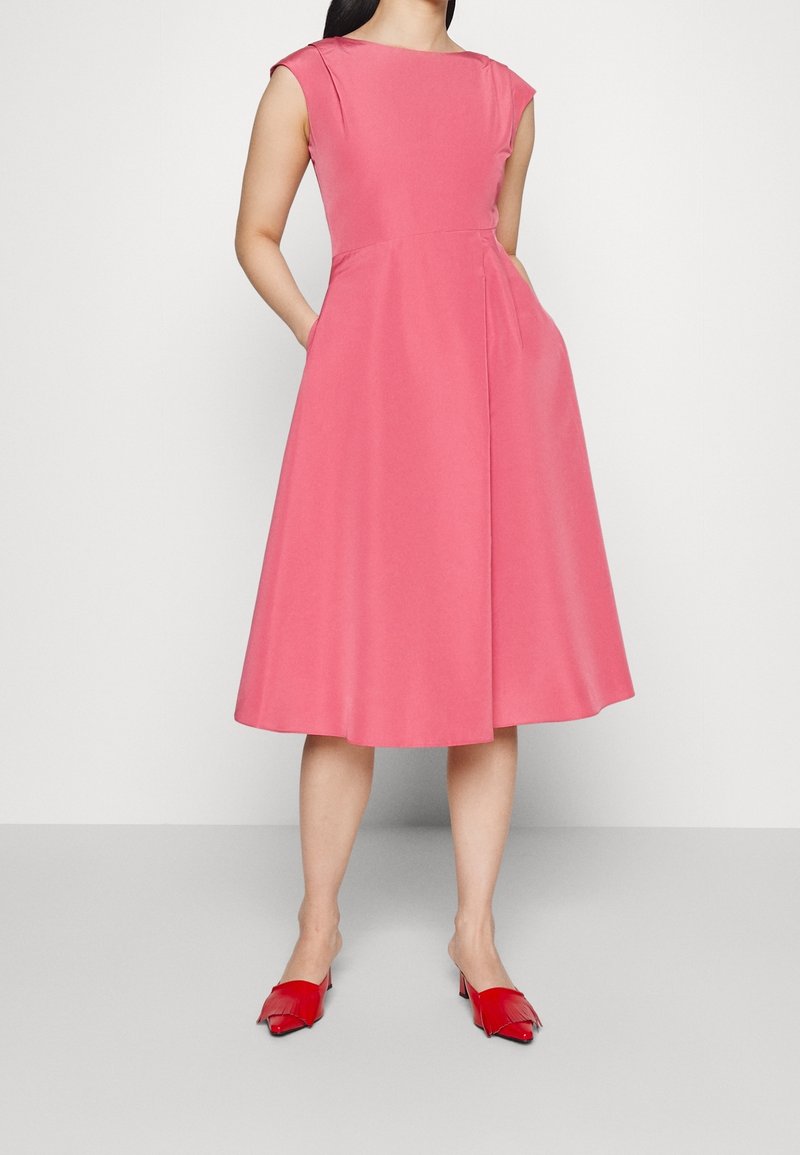 Femme portant une robe rose sans manches, longueur genoux, avec des poches, et des chaussures rouges à bouts pointus à talons bas, debout devant un fond uni.
