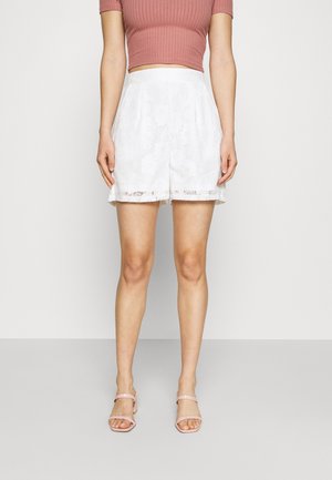 Femme portant un short en dentelle blanche taille haute, un top côtelé court rose et des sandales à talons roses clairs à brides, debout devant un fond blanc.