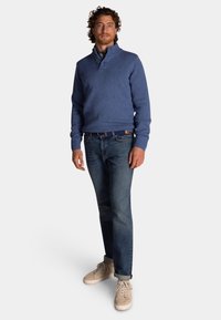 Blauwe gebreide trui met een hoge kraag, voorzien van drie knoopaccenten. Gepaarde met donkerblauwe jeans en beige sneakers.