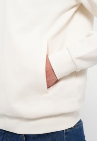 Felpa crema con scollatura rotonda, polsini a coste e tasca laterale. Texture liscia e design semplice; anche il bordo in vita è a coste.