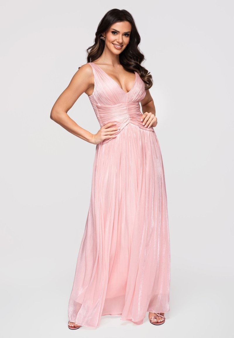 Femme souriante portant une robe de soirée rose fluide sans manches, debout les mains sur les hanches devant un fond uni.