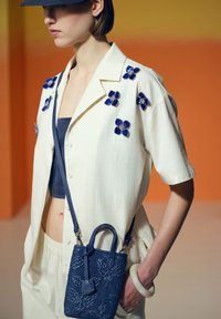 Camisa blanca de manga corta con bordado floral azul, bolso cruzado azul marino con diseño texturizado y una pulsera grande translúcida.