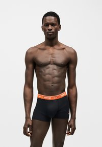 Homme athlétique portant un boxer noir Nike avec une ceinture orange, debout devant un fond blanc uni, regardant devant lui.