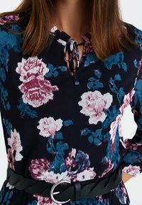 Robe fleurie en tissu bleu marine foncé, ornée de grandes fleurs roses et turquoise, avec un col arrondi à nouer et une ceinture tressée noire à boucle argentée.