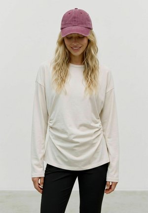 Femme blonde portant une chemise blanche à manches longues, un pantalon noir et une casquette de baseball rose, regardant vers le bas et souriant sur un fond uni.
