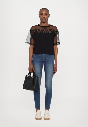 Schwarzes Oberteil mit transparenten Ärmeln und gemustertem Oberteil, kombiniert mit engen blauen Jeans und einer kleinen schwarzen Handtasche. Neutrale Sneaker vervollständigen das Outfit.
