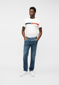 Άνδρας με λευκό γραφικό μπλουζάκι, μπλε τζιν και λευκά αθλητικά παπούτσια, στέκεται με τα χέρια στις τσέπες, μπροστά σε απλό φόντο.