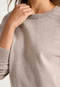 Helles beiges Sweatshirt mit geripptem Halsausschnitt und Bündchen, das eine weiche Textur und eine lässige Passform bietet. Hände berühren den Kragenbereich.