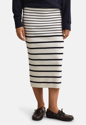 Person iført en hvid midi-nederdel med sorte horisontale striber, en mørk langærmet top og mørkebrune snøreløse loafers, stående.