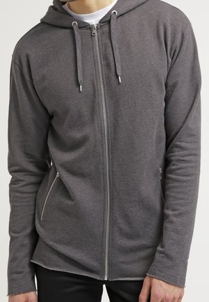 Mężczyzna ubrany w ciemnoszarą bluzę z kapturem na zamek, z troczkami i kieszeniami na zamek, w połączeniu z czarnymi spodniami i białą koszulką wewnętrzną.