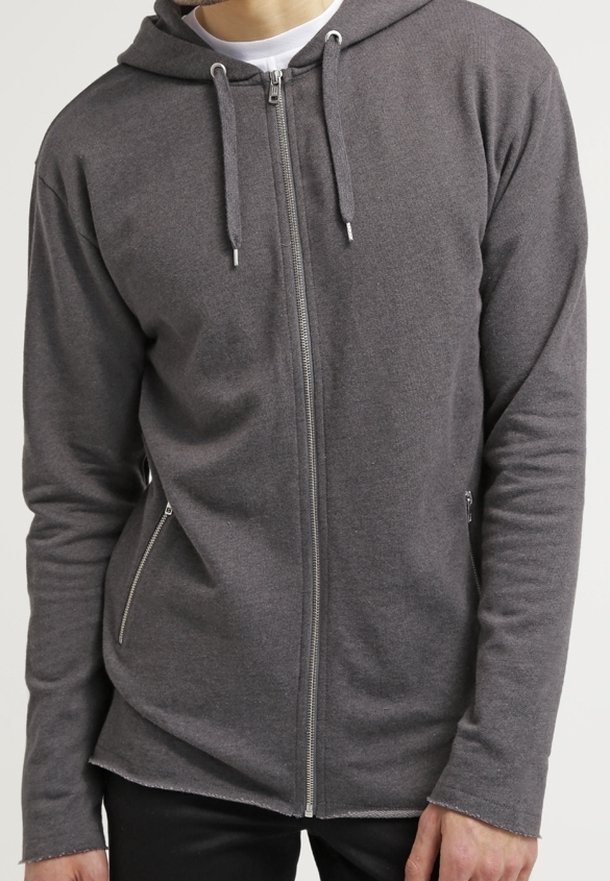 Homme portant un sweat à capuche zippé gris foncé avec cordons de serrage et poches zippées, associé à un pantalon noir et un t-shirt blanc en dessous.