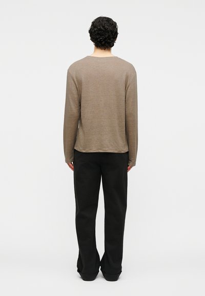 Άτομο με σγουρά μαλλιά που στέκεται με την πλάτη γυρισμένη, φοράει ανοιχτό καφέ μπλούζα μακρυμάνικη και μαύρο παντελόνι με φαρδιά πόδια, σε απλό φόντο.