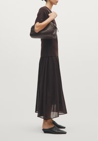 Sac à main en tricot marron avec un design texturé, porté par un mannequin avec un haut marron et une jupe longue, fluide et semi-transparente. Chaussures noires à enfiler.