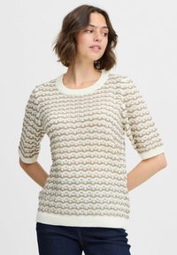 Suéter de manga corta en crema y beige con un patrón de olas texturizadas, cuello y puños de canalé. Se combina con denim oscuro.