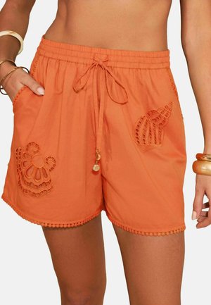 Orangefarbene Shorts mit Kordelzug, mit Muschel- und Wellen-Applikationsstickerei, getragen mit goldenen Armreifen am rechten Handgelenk und Armbändern am linken Arm.