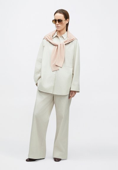 Chemise ample beige clair, pantalon large assorti, et un pull rose pâle noué autour du cou. Lunettes de soleil marron au design simple.