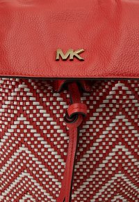 Sac à main rouge tissé avec des accents blancs, doté d'un rabat en cuir rouge lisse, d'un logo "MK" doré, et d'une bandoulière réglable avec un détail pompon.