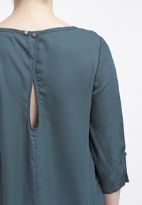 Blouse légère teal à manches trois quarts, avec une ouverture en forme de clé au dos et deux boutons-pression au cou. Texture lisse.