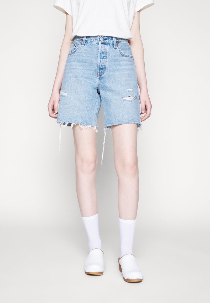 Personne portant un short en jean déchiré bleu clair, des chaussettes blanches, des sabots blancs sans lacets et une chemise blanche à manches courtes sur un fond uni.