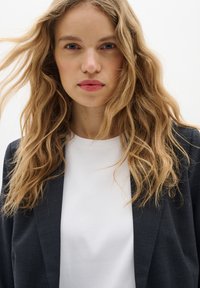 Černý blazer přes bílé tričko; modelka má dlouhé, vlnité blond vlasy; neutrální pozadí; zaměření na obličej a horní část těla.