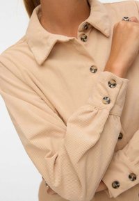 Blusa in corduroy beige con colletto, chiusura con bottoni e maniche a sbuffo con cuciture visibili e dettagli in metallo sui bottoni.