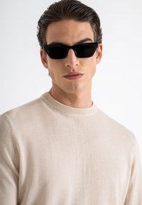 Occhiali da sole neri a forma di occhio di gatto indossati con un maglione girocollo beige chiaro, che mette in evidenza la superficie liscia del tessuto e le linee pulite.