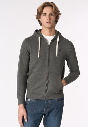 Giovane uomo che indossa una felpa grigia scura con zip e cordini bianchi e jeans blu, in piedi con una mano in tasca contro uno sfondo semplice.