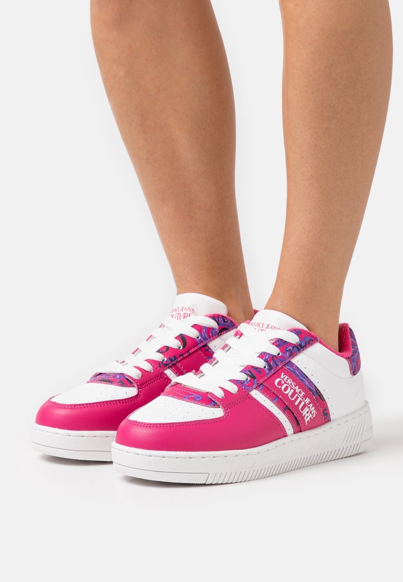Baskets basses blanches et roses avec des accents violets, des lacets blancs et le logo Versace Jeans Couture sur le côté.