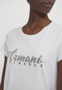 Camiseta blanca de algodón con "Armani Exchange" en pedrería negra, mangas cortas y cuello redondeado. Textura suave.