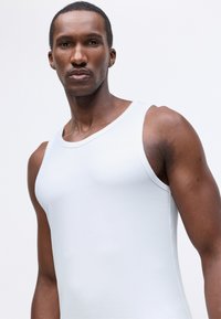 Balta kokvilnas bez piedurknēm blūze ar klasisku dizainu, gludu tekstūru un apaļu kakla izgriezumu, parādīta uz modeļa pret gaišu fonu.