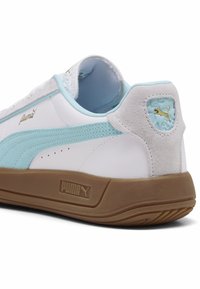 Sneaker Puma cu un superior din piele albă, accentuat cu suede albastru deschis, o talpă din cauciuc și detalii cu logo-uri aurii pe lateral și călcâi.