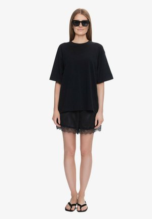 Kobieta w oversize czarnej koszulce, czarnych szortach wykończonych koronką, czarnych klapkach i dużych czarnych okularach przeciwsłonecznych, stojąca na białym tle.