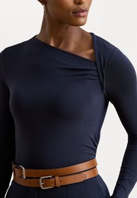 Marineblauw aansluitend langemouwen top met een gedrapeerde halslijn, gecombineerd met een brede bruine leren riem met een zilveren gesp. Het heeft een soepele textuur en een lichaamseigen ontwerp.