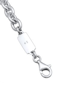 Sudraba ķēdes aproce ar lieliem posmiem, kurai ir taisnstūrveida birka ar iezīmi 925 un aizdare ar omāra aizdari drošai aizvēršanai.