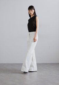 Schwarzes ärmelloses Oberteil kombiniert mit hochgeschnittenen, weißen, weit geschnittenen Hosen, die eine glatte Textur und eine klassische Silhouette aufweisen. Einfache schwarze Schuhe.