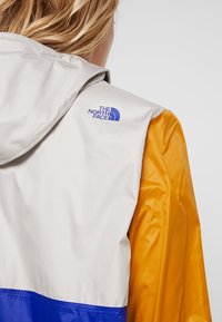 Färgblockerad vindjacka med en grå huva, orangea ärmar och en blå detalj vid nederkanten. Visar "The North Face"-logotypen på axeln.