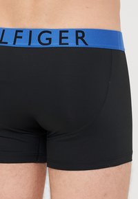 Schwarze Boxer-Shorts mit einem blauen elastischen Bündchen, auf dem der Markenname in fetten schwarzen Buchstaben steht. Glatte Textur, enganliegendes Design.