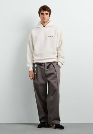 Junger Mann trägt einen weißen Fleece-Hoodie, lockere graue Hose mit Schlüsselanhänger und schwarze Schuhe, steht vor einer schlichten weißen Wand.