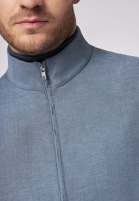 Hellblaues Reißverschluss-Jacke mit hohem Kragen und einem gestrickten marineblauen Akzent, glatter Textur und dezenten Steppnähten entlang des Reißverschlusses.